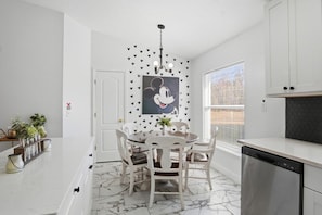 A kitchen with white cabinets, a marble floor, a dining table with chairs, a dishwasher, and a window with blinds.