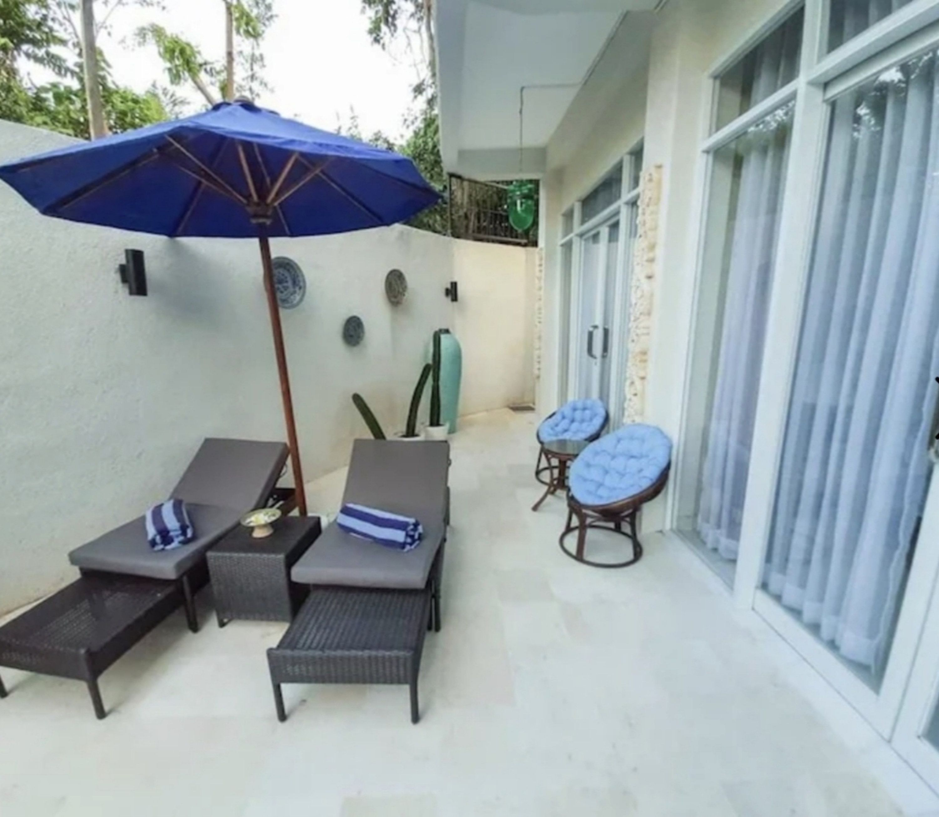 Um pátio com duas cadeiras de descanso, um guarda-sol azul e uma mesa com um objeto decorativo.