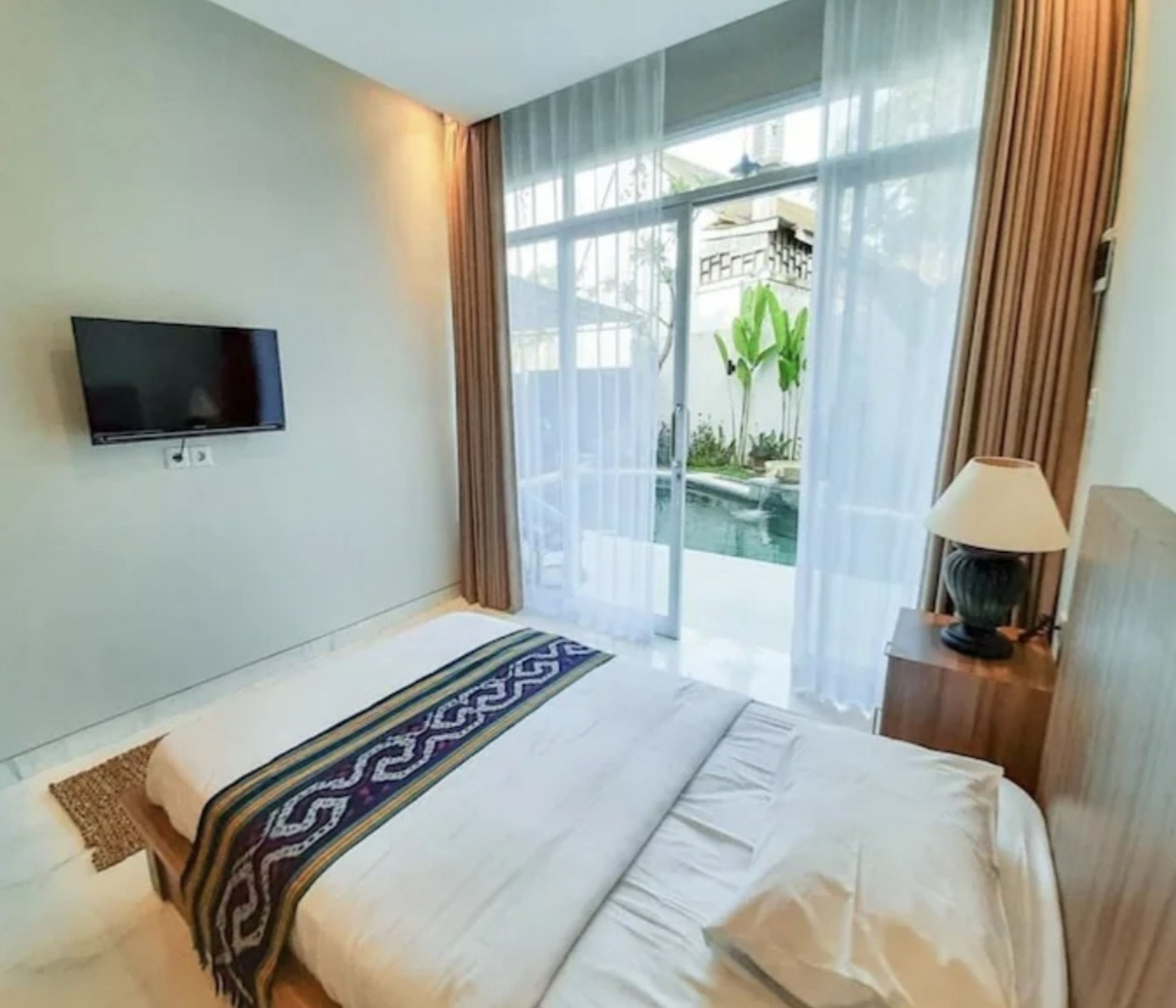 Habitación de hotel con cama, televisor montado en la pared, lámpara y vista a una piscina a través de un ventanal.