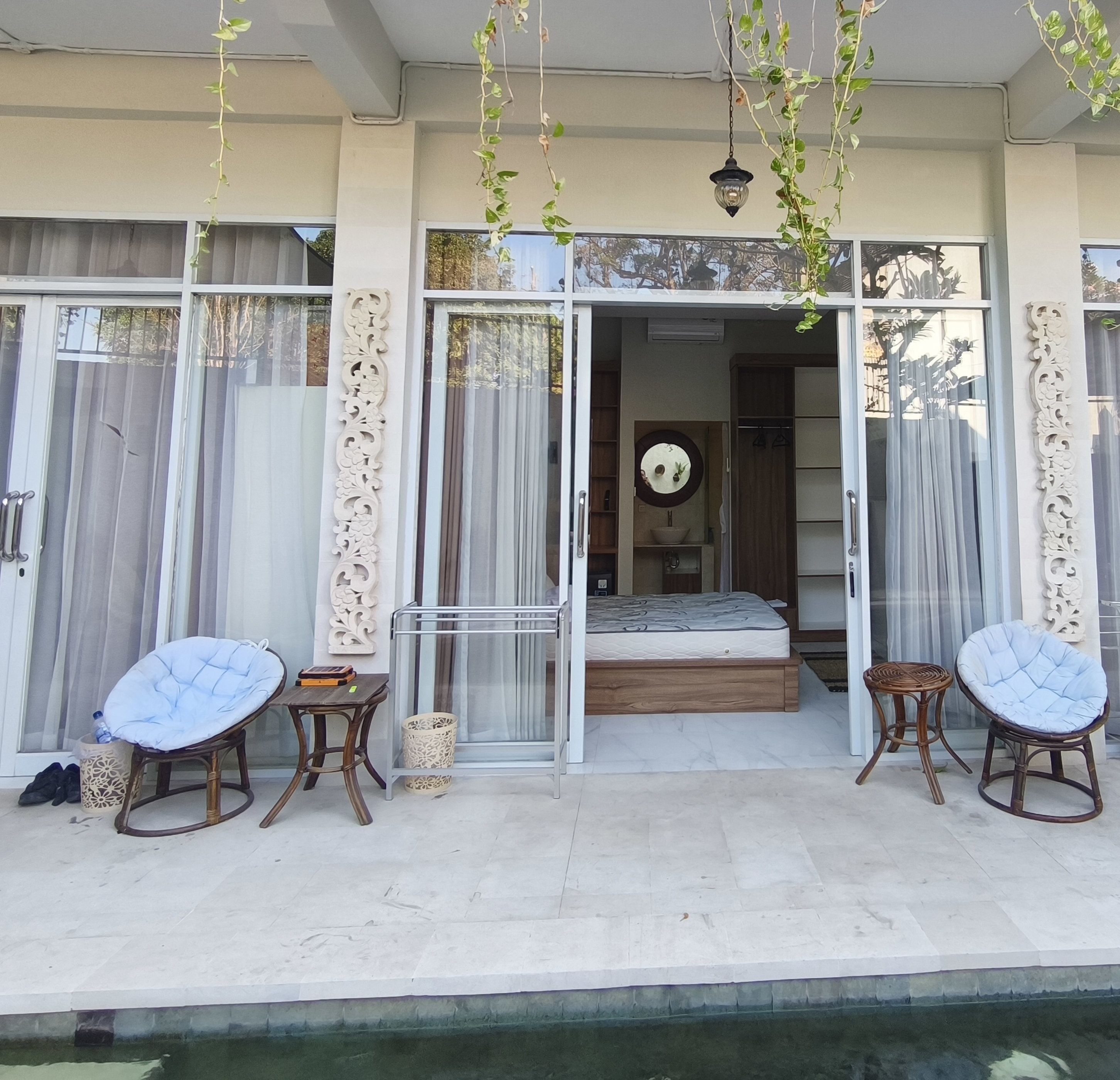 Un área de estar moderna al aire libre con dos sillones acolchados y una mesita, una entrada con puerta de vidrio a un dormitorio y vista a la piscina.