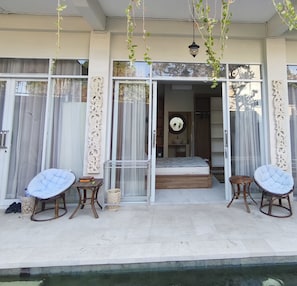 A modern outdoor seating area with two cushioned chairs and a small table, a glass door entrance to a bedroom, and a poolside view. - OYO 95416 Griya House Bstc (Denpasar)