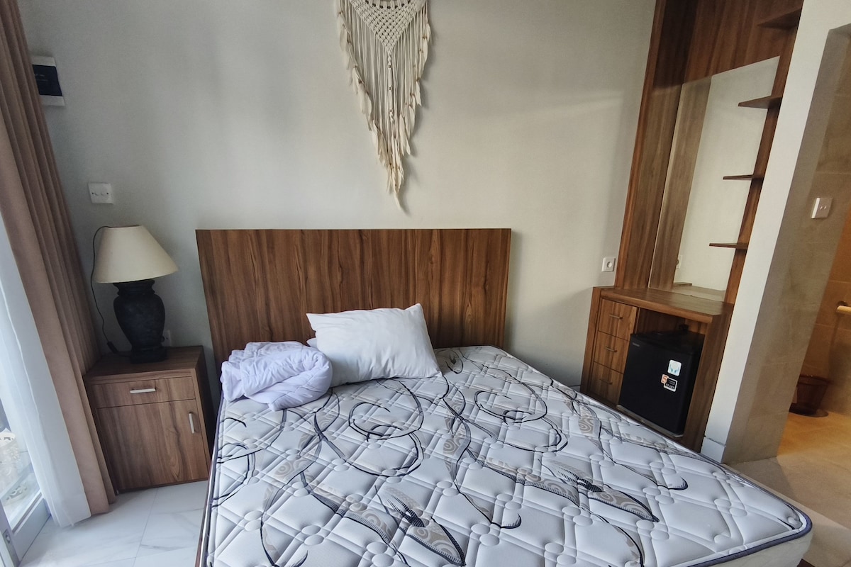 A bedroom with a wooden headboard, a bed with a patterned mattress, a bedside table with a lamp, and a wall-mounted macrame decoration.