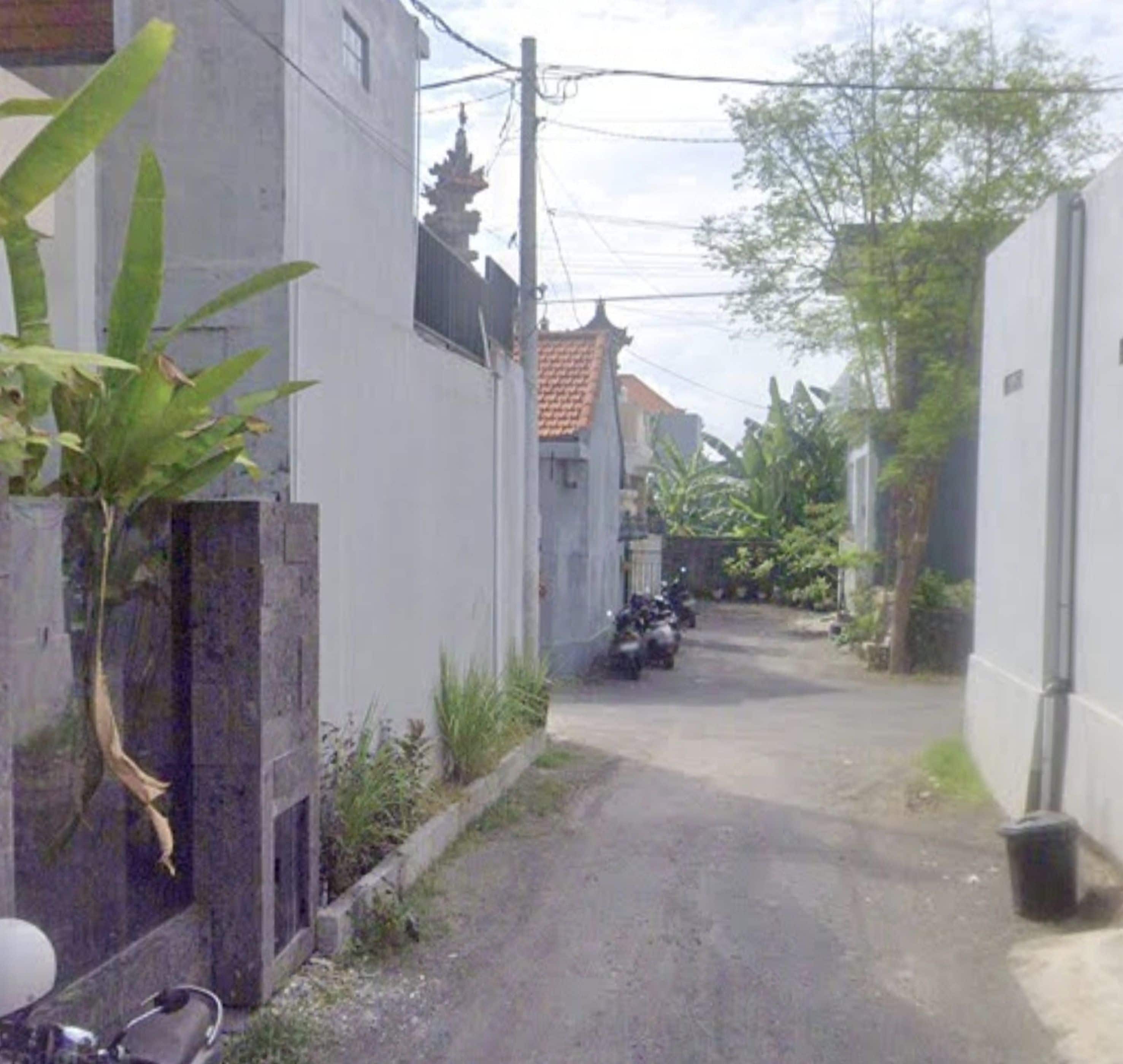 Uma rua estreita com motocicletas estacionadas, um edifício branco com telhado em azulejos e um muro de concreto com uma planta.