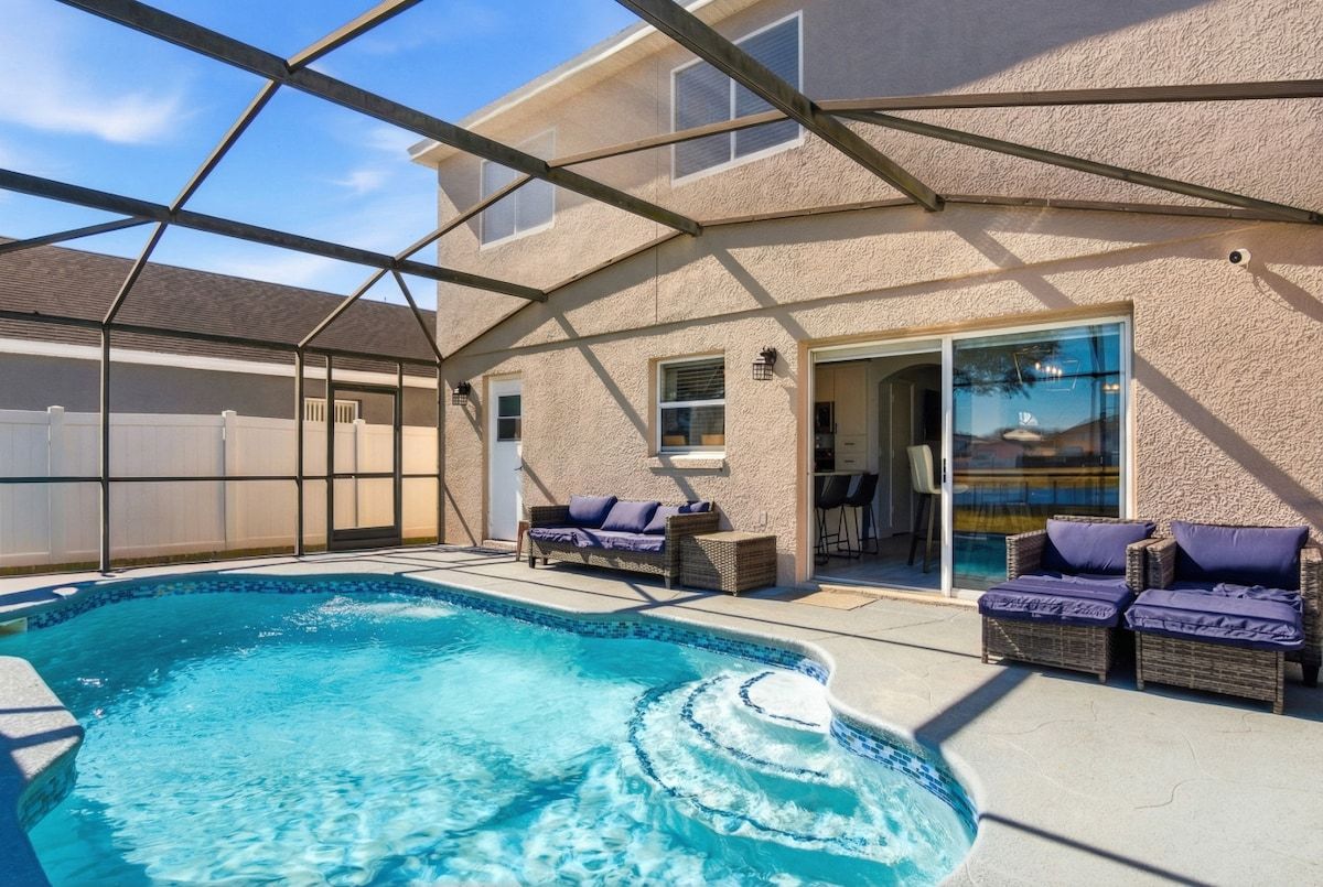 A covered outdoor swimming pool with lounge chairs, a patio area, and a house in the background.