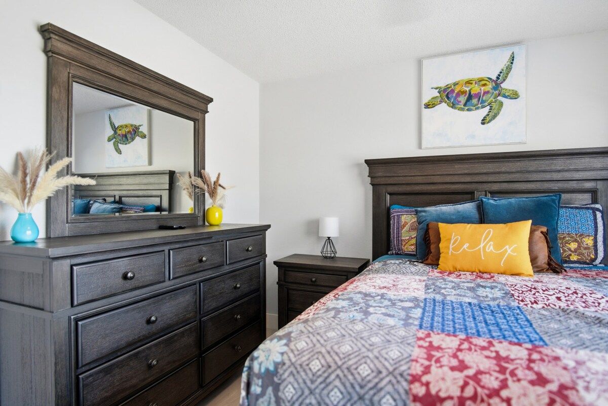 Ein Schlafzimmer mit dunklem Holzschrank, einem Bett mit gemusterten Textilien, einer Kopfteile mit Kissen, einem Wandspiegel und einem gerahmten Kunstwerk mit einer Schildkröte.