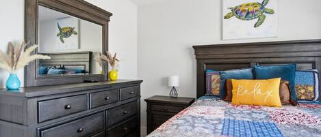 A bedroom with a dark wooden dresser, a bed with patterned bedding, a headboard with pillows, a wall-mounted mirror, and a framed artwork of a turtle.