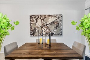 Un coin repas comprenant une table en bois, deux chaises, un tableau abstrait accroché au mur et deux plantes en pot.