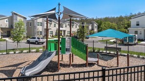 A playground with a slide and climbing structures, surrounded by a fence, with buildings in the background. - King Bed | Disney Contemporary Townhouse (Kissimmee)