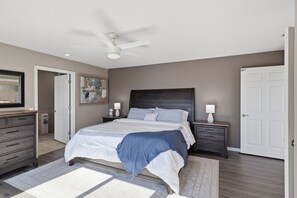 A bedroom with a bed, bedside tables, a ceiling fan, a door, and a painting on the wall. - Lakefront Paradise with 4 Decks and Unmatched Panoramic Views! (Sunrise Beach)