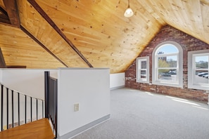A spacious attic room with a sloped wooden ceiling, a staircase with metal railings, and large windows offering a view of a parking lot and buildings.