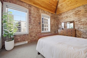 A bedroom with a brick wall, a wooden ceiling, a large bed, a dresser, and a potted plant.