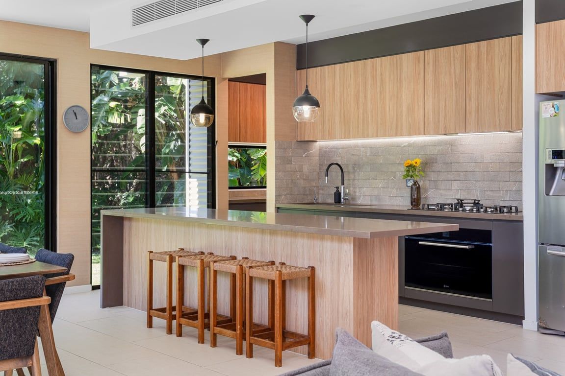A modern kitchen with a central island, bar stools, and a view of greenery outside.
