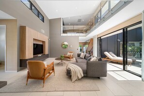 A modern living room with a sofa, armchair, and TV, a staircase, and a balcony with sun loungers.