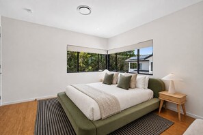 A modern bedroom with a large bed, a nightstand, and a view of greenery outside.