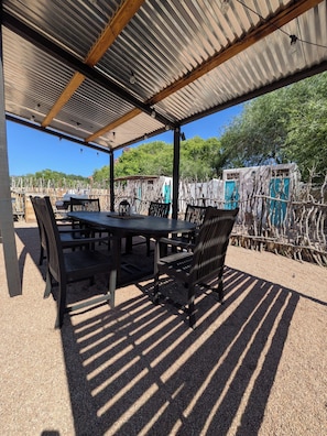Une terrasse extérieure couverte, avec une table et des chaises, entourée d’un sol en gravier et d’une clôture en métal.