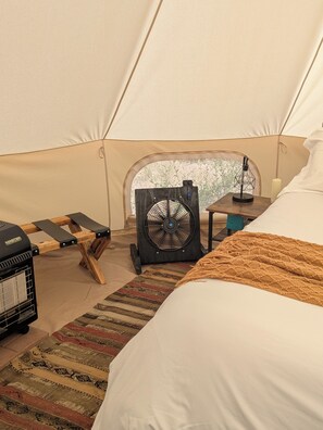 A tent interior with a bed, a fan, a heater, and a table with a lamp.