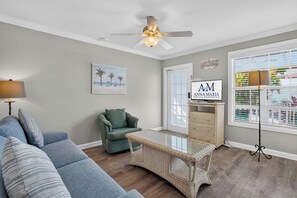 A living room with a sofa, a chair, a glass-top coffee table, a ceiling fan, a beach-themed painting, and a sign for Anna Maria Rental Properties. - Tradewinds Cottage 11 | Steps to Beach • Bayfront Heated Pool (Bradenton Beach)