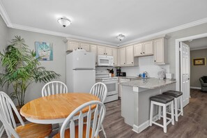 A modern kitchen with white cabinets, a wooden dining table, and a built-in island with bar stools. - Tradewinds Cottage 11 | Steps to Beach • Bayfront Heated Pool (Bradenton Beach)