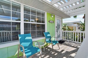 A covered porch with two chairs and a table, a green wall, and a white door. - Tradewinds Cottage 11 | Steps to Beach • Bayfront Heated Pool (Bradenton Beach)