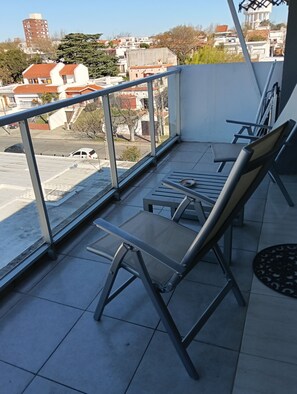 A balcony with a view of a cityscape, including buildings and a road. There are two lounge chairs and a small table.