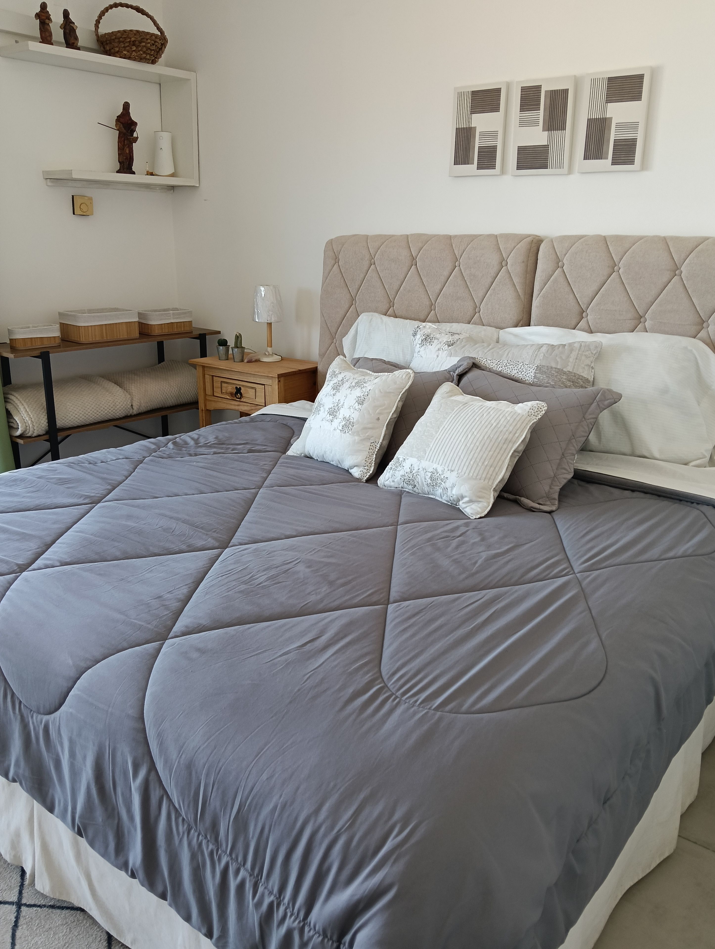 A neatly made bed with a grey quilt, a bedside table with a lamp, and a shelf with decorative items.
