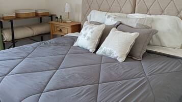 A neatly made bed with a grey quilt, a bedside table with a lamp, and a shelf with decorative items.