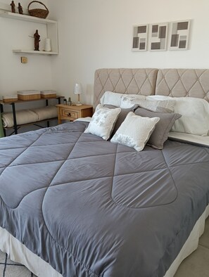 A neatly made bed with a grey quilt, a bedside table with a lamp, and a shelf with decorative items.