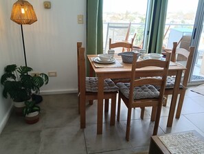 A dining area with a table set for two, wooden chairs with cushions, a floor lamp, and potted plants.