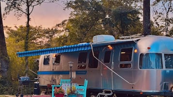 Una caravana antigua con un toldo azul, una pequeña mesa con sillas y plantas en macetas.