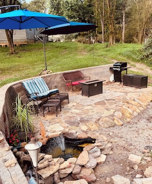 A stone patio with outdoor furniture, a small water feature, and a blue umbrella.
