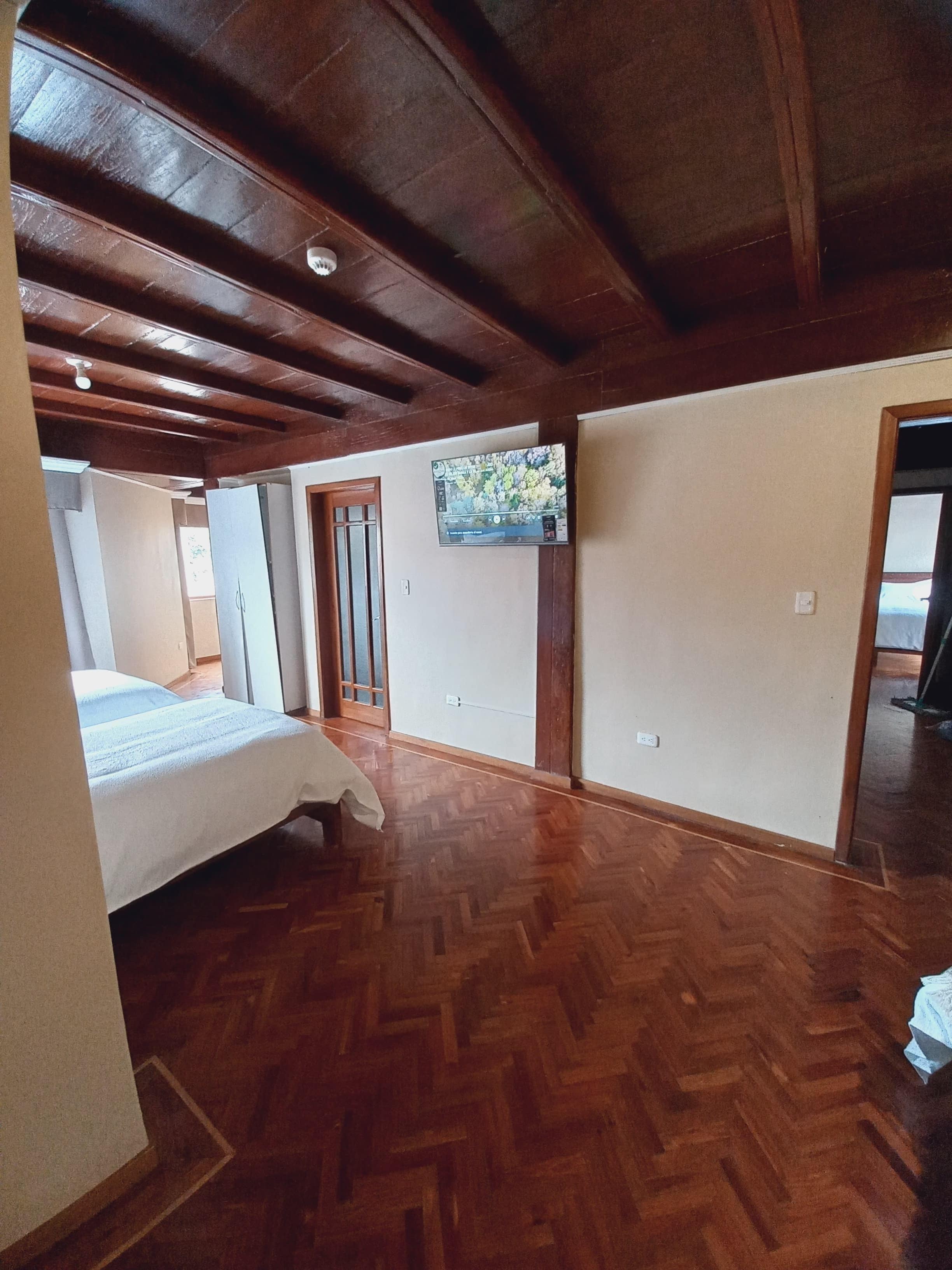 Una habitación con suelo de madera en patrón de espiga, una cama, un televisor en la pared y una puerta que da a otra habitación.