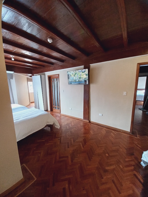 Una habitación con suelo de madera en patrón de espiga, una cama, un televisor en la pared y una puerta que da a otra habitación.