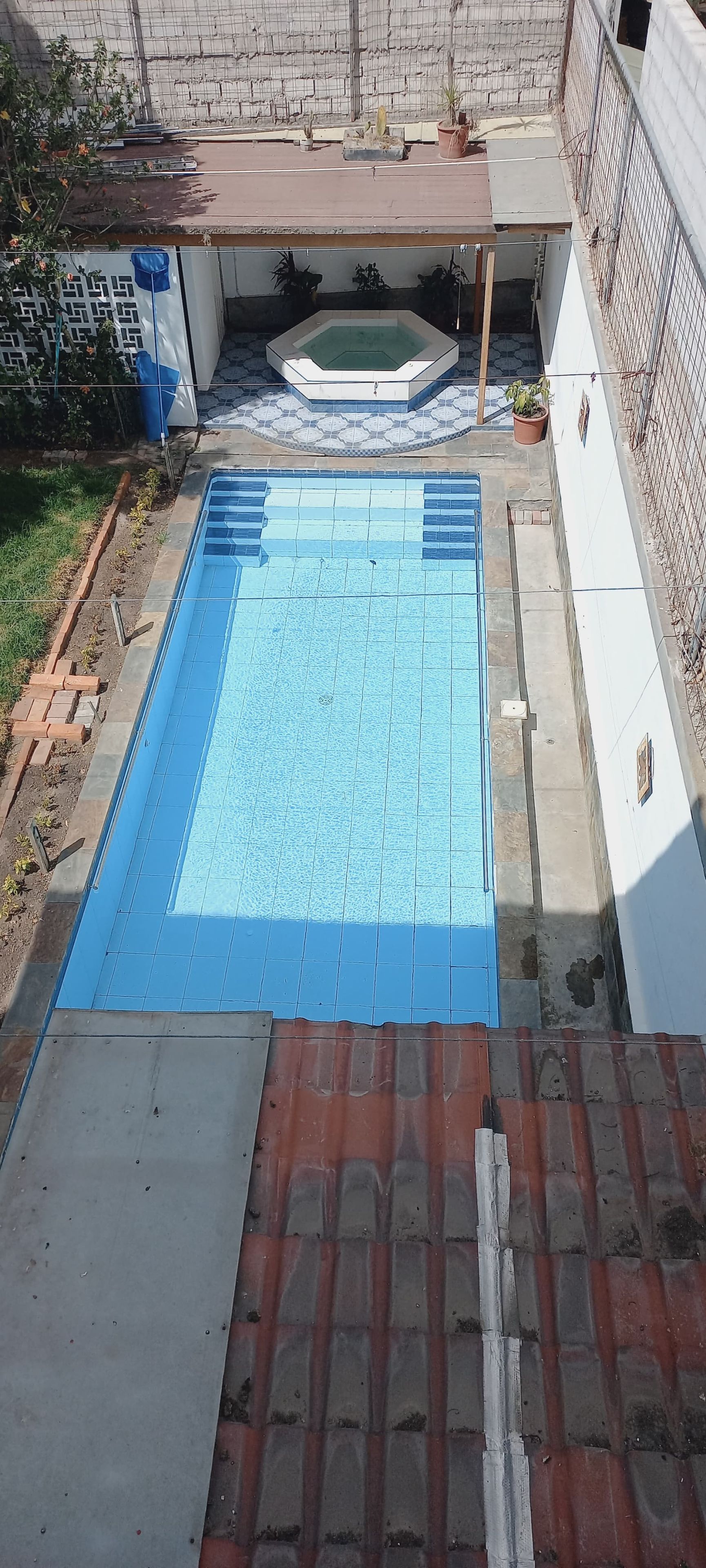 Una piscina con zona ajardinada empavedada, rodeada por un muro de ladrillo y una cerca de alambrada.