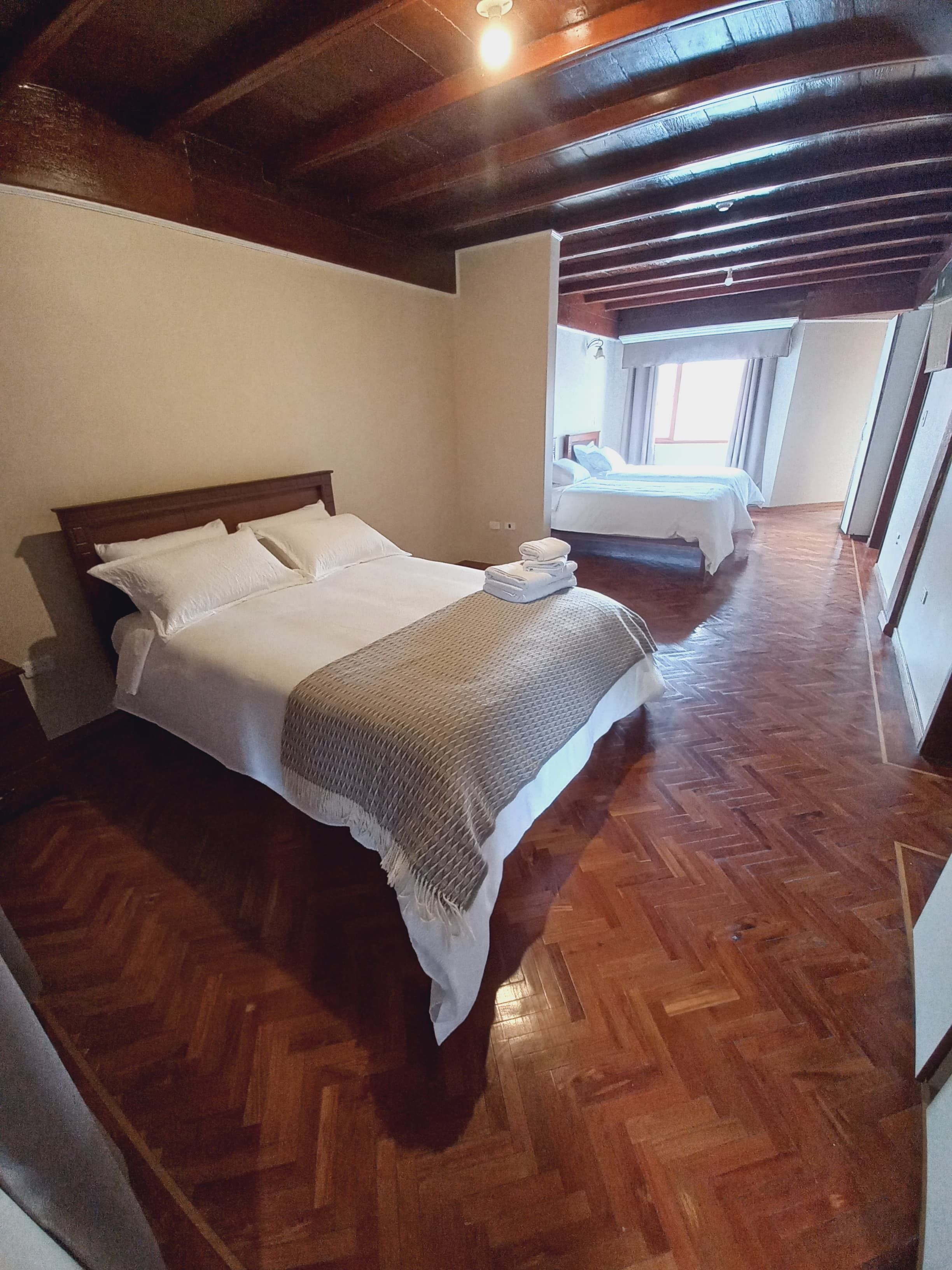 A hotel room with a wooden floor, a bed with white linens, and a view of another room through an open door.