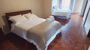 A hotel room with a wooden floor, a bed with white linens, and a view of another room through an open door.