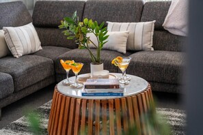 A round wooden coffee table with a white marble top, surrounded by a grey sofa with striped cushions, a potted plant, and two cocktail glasses with orange slices. - Cosy Corner on Macdonell (Taupo)