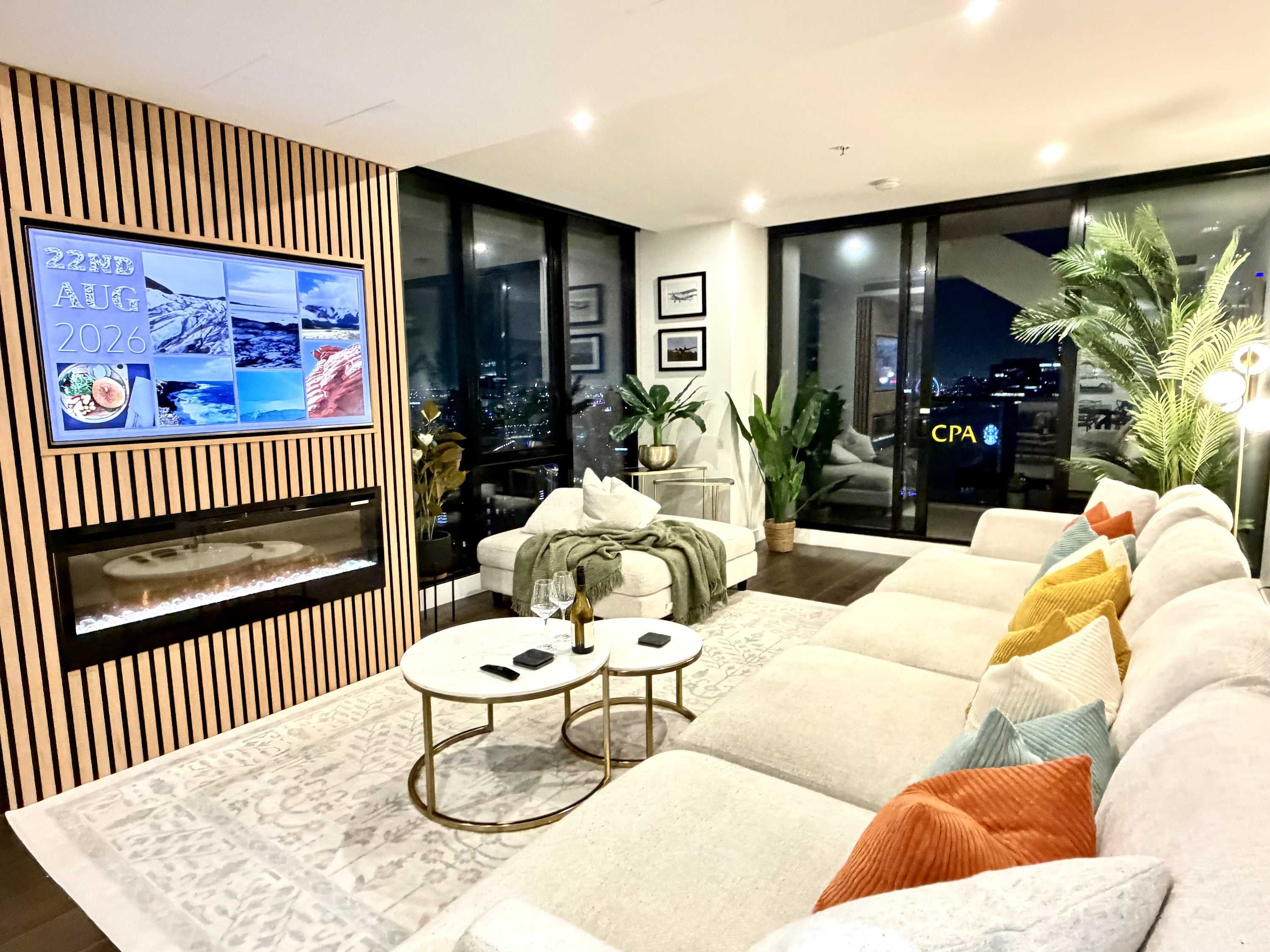 A modern living room with a fireplace, a television mounted on a wooden panel, a sofa set, and a view of the city at night.