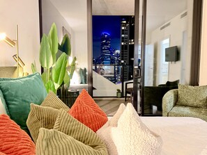 A modern hotel room with a city view, a sofa, a bed with colorful pillows, and a plant. - 36th Floor Luxury 2BR City Views Pool & 2 Cars (Southbank)