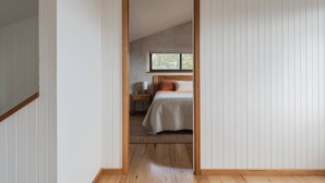 Une chambre avec un plafond en pente, un lit recouvert d’une couette blanche, une table de chevet avec une lampe et une fenêtre donnant sur des arbres.