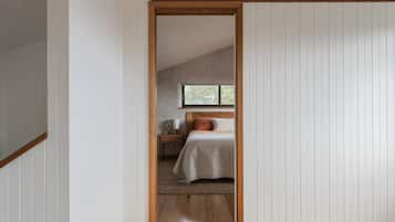 Une chambre avec un plafond en pente, un lit recouvert d’une couette blanche, une table de chevet avec une lampe et une fenêtre donnant sur des arbres.