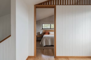Ein Schlafzimmer mit schrägem Dach, einem Bett mit weißer Daunendecke, einem Nachttisch mit Lampe und einem Fenster mit Blick auf Bäume.