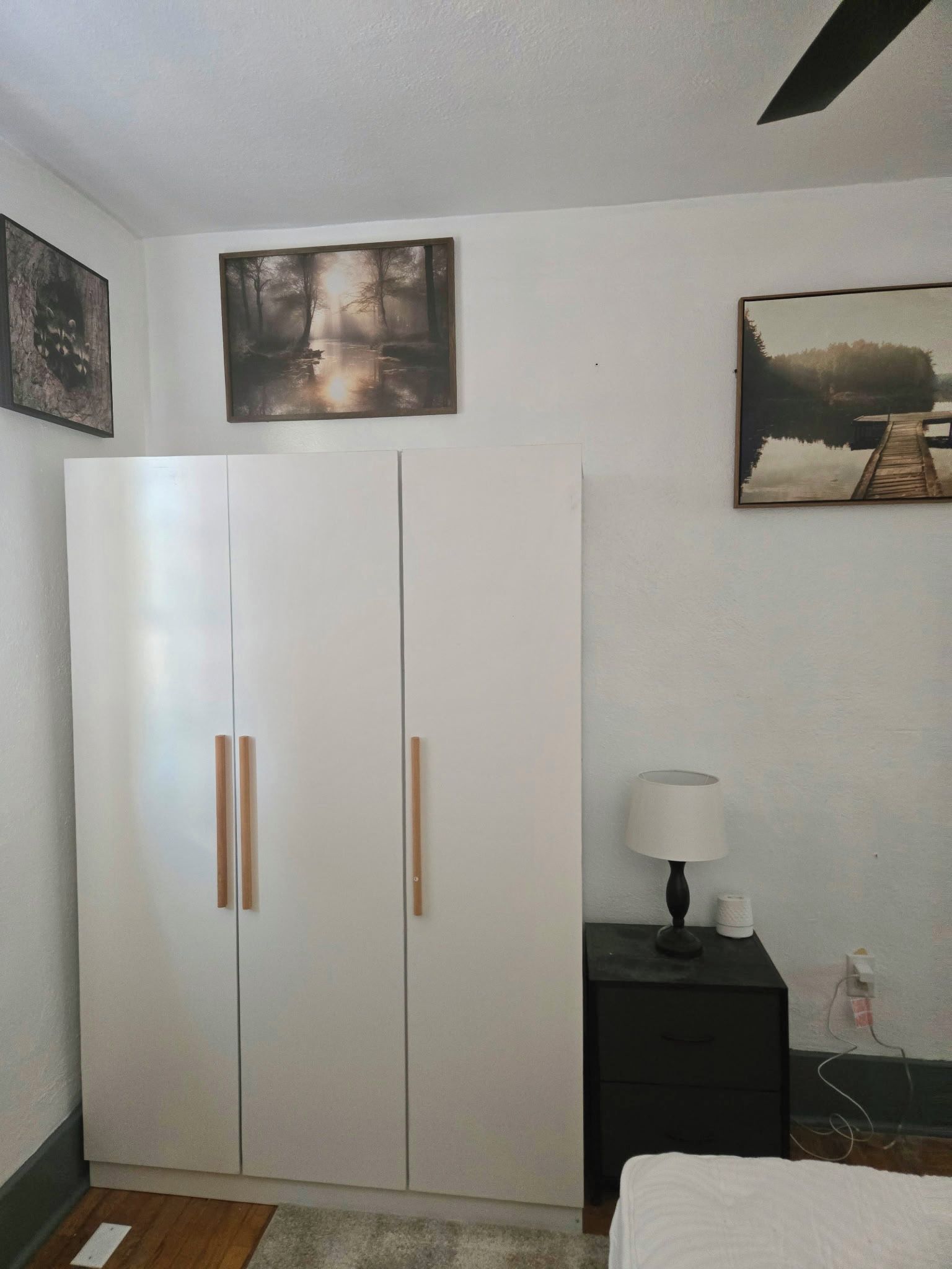 A bedroom with a wardrobe, a bedside table with a lamp, and two framed pictures on the wall.