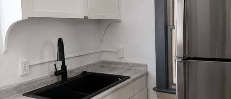 Una cocina con armarios blancos, un lavamanos negro, un refrigerador de acero inoxidable y una ventana con marco oscuro.