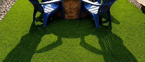A circular fire pit surrounded by four blue chairs, set on a patch of artificial grass.