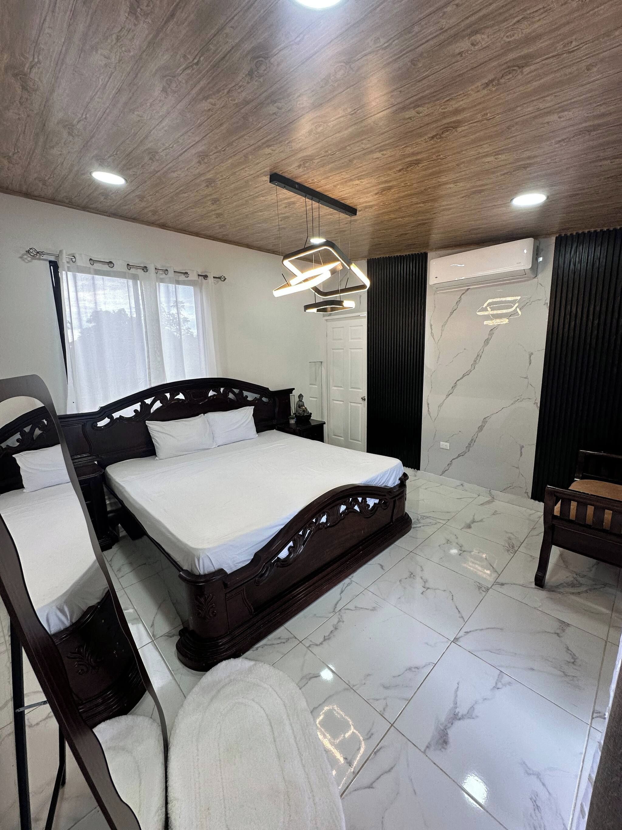 Une chambre à coucher avec un lit doté d’une tête de lit en bois, une table de chevet, une chaise, un miroir, un luminaire au plafond, un climatiseur fixé au mur et une fenêtre avec des rideaux.