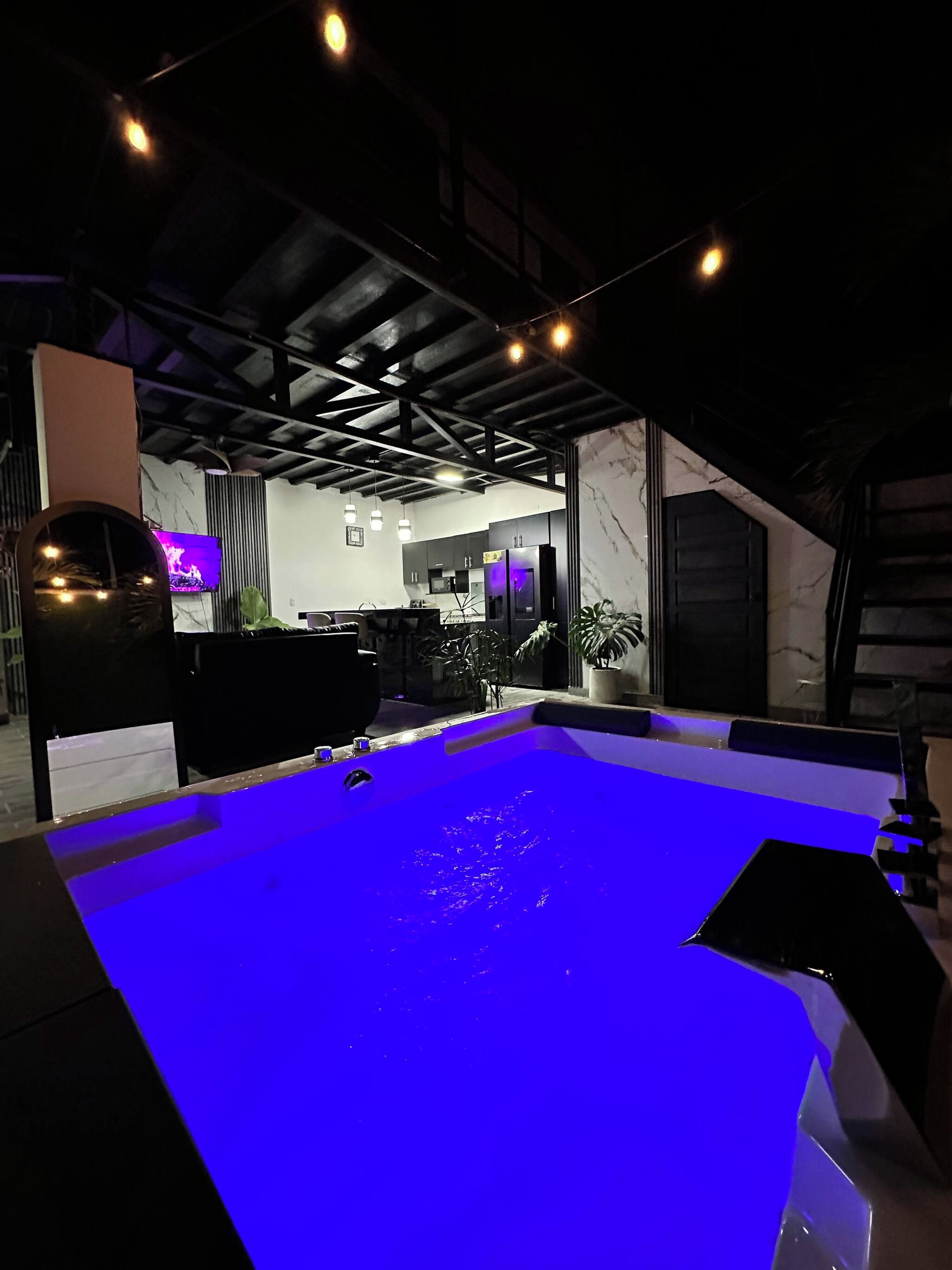 Zona de piscina con superficie de agua iluminada en azul, una cocina al fondo y techo con vigas a la vista.