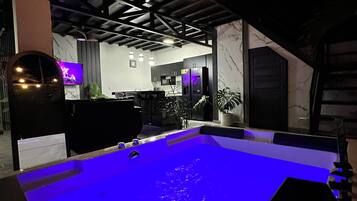 Zona de piscina con superficie de agua iluminada en azul, una cocina al fondo y techo con vigas a la vista.