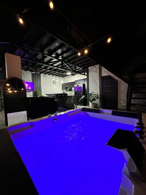 A pool area with a blue-lit water surface, a kitchen in the background, and a ceiling with exposed beams. - Betandy Apartment Jacuzzi (Rita)