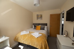 A hotel room with a bed, a bedside table, a mirror, and a television on the wall. - Modern 4-Bed House | Windsor | Driveway + Garden (Windsor)