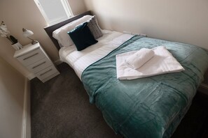 Een netjes opgemaakt bed met een blauwgroene dekbedovertrek, witte kussens en een opgevouwen witte handdoek.